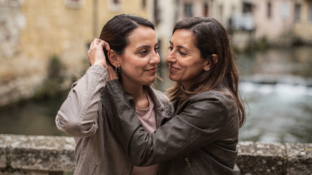 Deux amies sur un pont de pierre au-dessus de l’eau, l’une relève ses cheveux pour montrer sa créole à l’autre