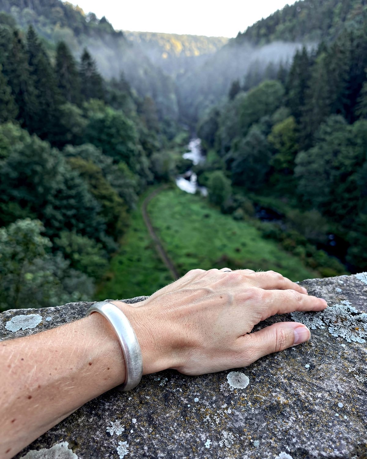 Un jonc en argent sterling 925 au poignet, la main posée sur le parapet de pierre d’un pont, une vallée boisée et encaissée en contrebas